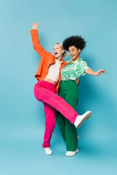 Full length of stylish african american woman smiling at camera near excited friend screaming on blue background — Stock Photo