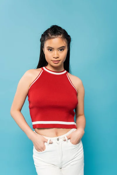 Brunette asian woman in red top looking at camera while posing with hands in pockets isolated on blue — Stock Photo