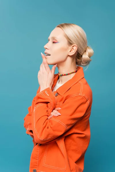 Side view of blonde woman in orange jacket and colorful beads posing with hand near chin isolated on blue — Stock Photo