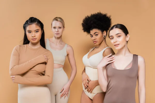 Giovani e belle donne multiculturali guardando la fotocamera mentre posa in biancheria intima isolata sul beige — Foto stock