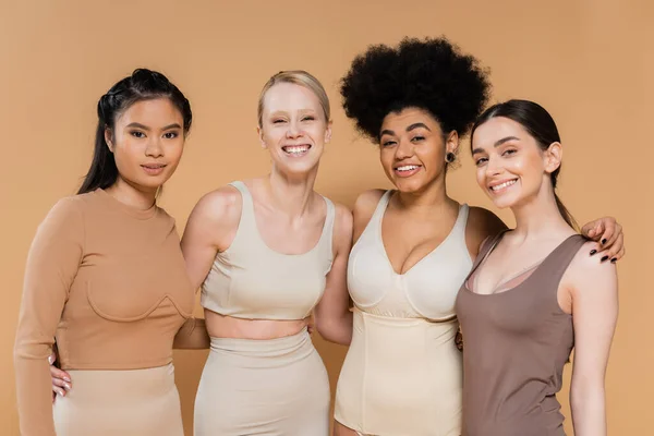 Cheerful multicultural women in lingerie embracing and looking at camera isolated on beige - foto de stock