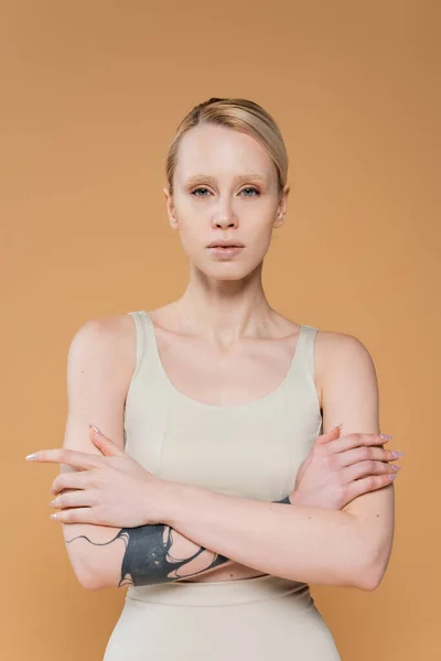 Front view of blonde tattooed woman in top posing with crossed arms and looking at camera isolated on beige — Stockfoto