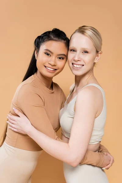 Blonde and brunette interracial women in underwear embracing and smiling at camera isolated on beige — Stockfoto