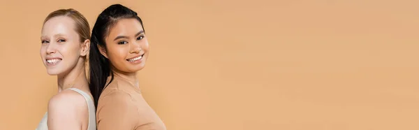 Cheerful multiethnic women posing back to back and looking at camera isolated on beige, banner — Stockfoto