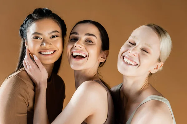 Jóvenes y felices mujeres multiculturales en ropa interior sonriendo a la cámara aislada en beige - foto de stock