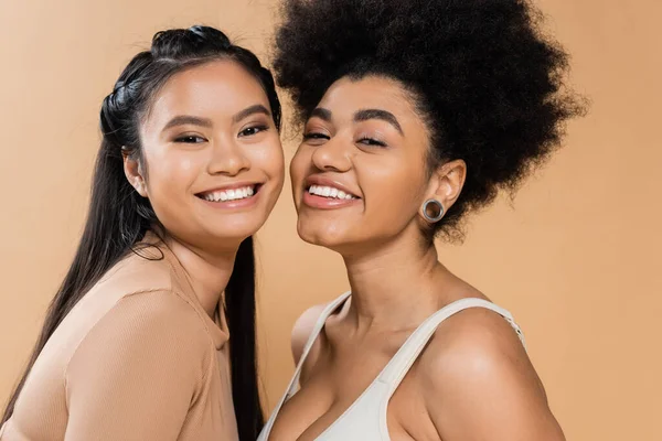 Portrait of pretty african american and asian women looking at camera isolated on beige - foto de stock