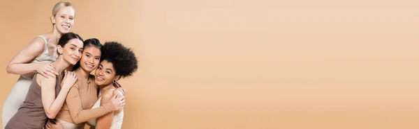 Young and happy multiethnic women in lingerie embracing and smiling at camera isolated on beige, banner — Stockfoto