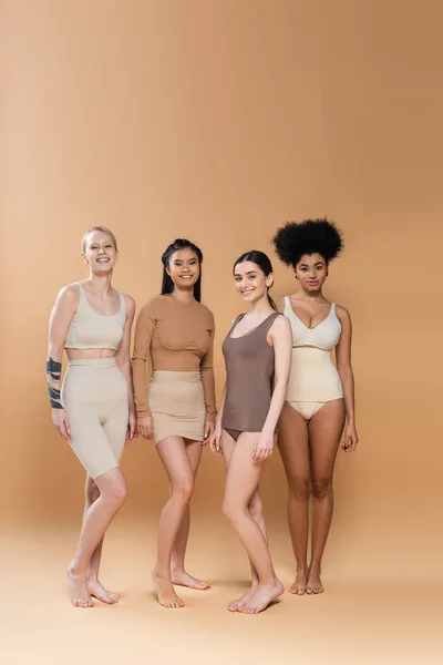 In voller Länge glückliche junge Frauen in Unterwäsche stehend und in die Kamera schauend auf beigem Hintergrund — Stockfoto