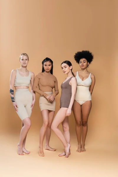 Полная длина босых многоэтнических женщин в нижнем белье, стоящих на бежевом фоне — стоковое фото
