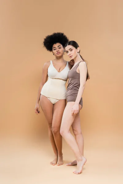 Longitud completa de las mujeres jóvenes multiétnicas posando en ropa interior sobre fondo beige - foto de stock