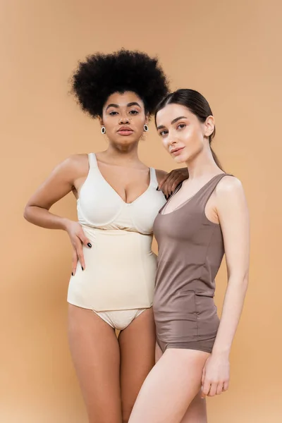 Pretty african american model in lingerie posing with hand on waist near young brunette woman isolated on beige — Fotografia de Stock