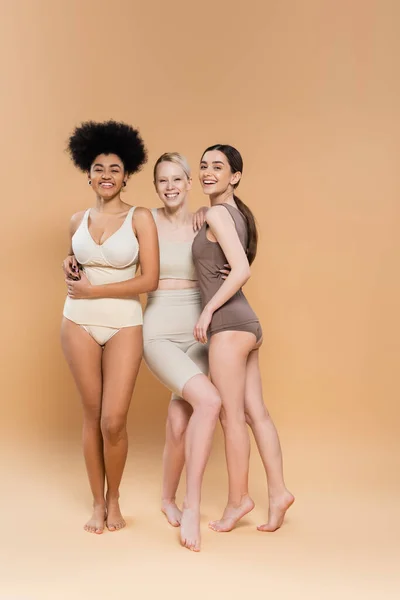 Longitud completa de modelos multiétnicos felices en lencería abrazando mientras está de pie sobre fondo beige - foto de stock