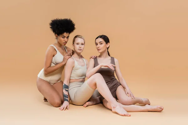 Full length of young multicultural women in underwear looking at camera while sitting on beige background — Stock Photo
