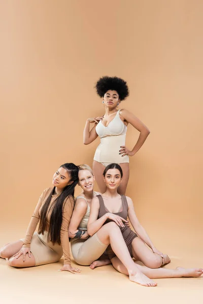 Full length of african american model in lingerie posing near young multiethnic women sitting on beige background - foto de stock