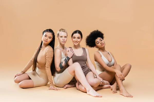 In voller Länge junge multiethnische Frauen in Unterwäsche sitzen und in die Kamera auf beigem Hintergrund schauen — Stockfoto