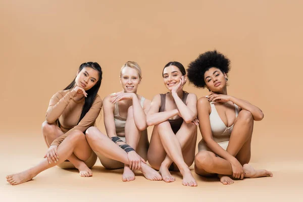 Full length of pretty multicultural models in underwear sitting with crossed legs on beige background — Stock Photo