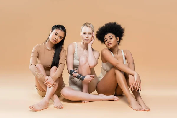Piena lunghezza di donne multiculturali a piedi nudi in biancheria intima seduto e guardando la fotocamera su sfondo beige — Foto stock