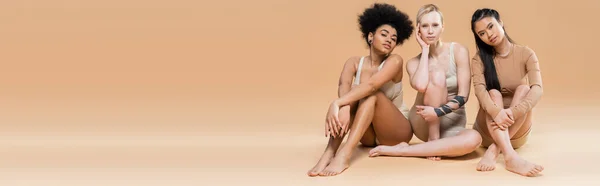 Full length of barefoot multiethnic women in lingerie looking at camera while sitting on beige background, banner — Fotografia de Stock