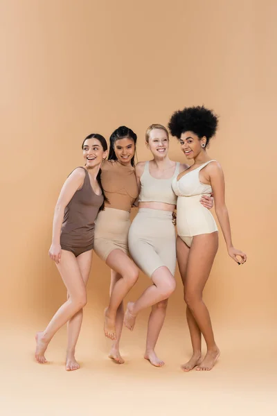 Full length of cheerful multicultural women in underwear embracing while standing on beige background — Foto stock