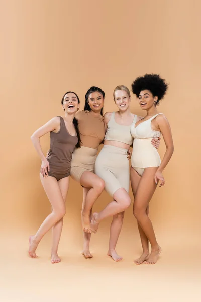 Piena lunghezza di giovani e felici donne multietniche in lingerie abbracciando e guardando la fotocamera su sfondo beige — Foto stock