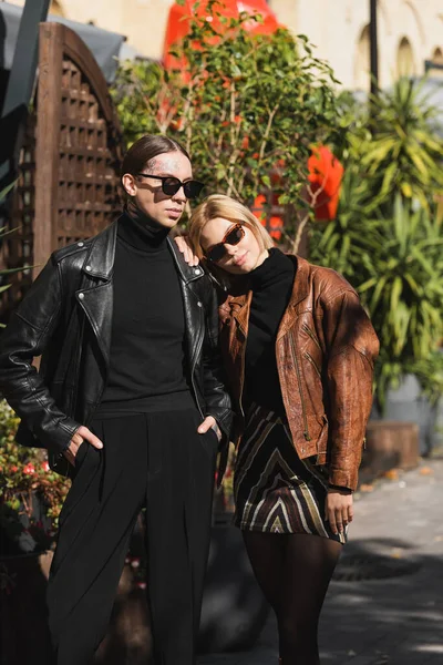 Blonde woman in sunglasses leaning on shoulder of stylish man posing with hands in pockets - foto de stock