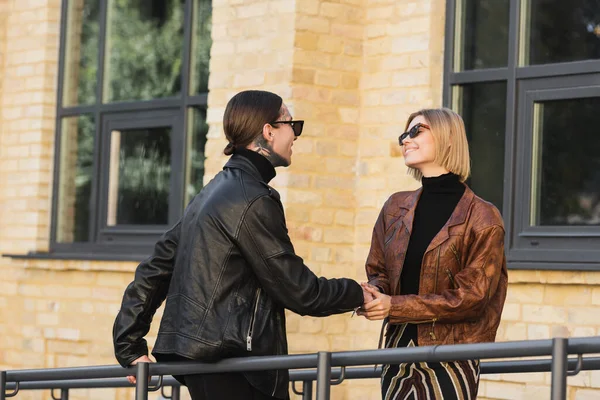 Couple élégant en lunettes de soleil et vestes en cuir souriant tout en se regardant et en se tenant la main — Photo de stock