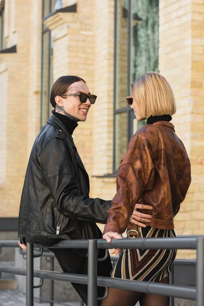 Happy tattooed man in stylish sunglasses and leather jacket hugging blonde girlfriend - foto de stock