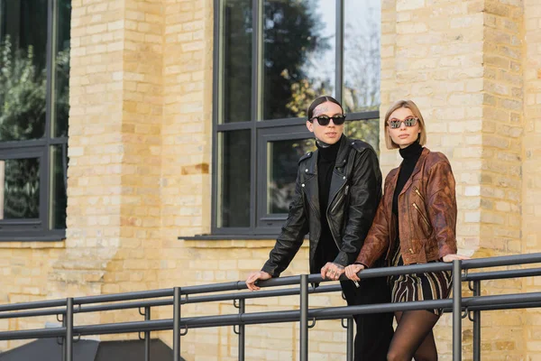 Young couple in trendy sunglasses and leather jackets standing together outdoors — Photo de stock