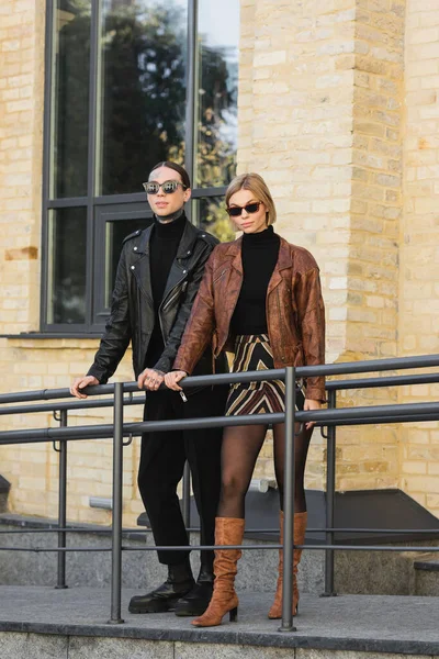 Comprimento total de elegante jovem casal em jaquetas de couro e óculos de sol em pé ao ar livre — Fotografia de Stock