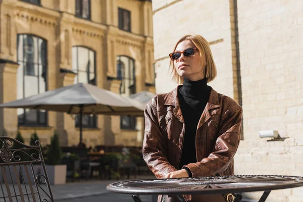 Blonde woman in sunglasses and stylish leather jacket sitting in outdoor cafe — Foto stock