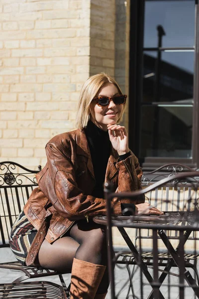 Mujer rubia feliz en gafas de sol y chaqueta de cuero elegante sentado en la cafetería al aire libre - foto de stock