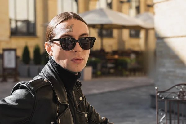 Tattooed young man in stylish sunglasses and black leather jacket looking at camera outdoors — Photo de stock