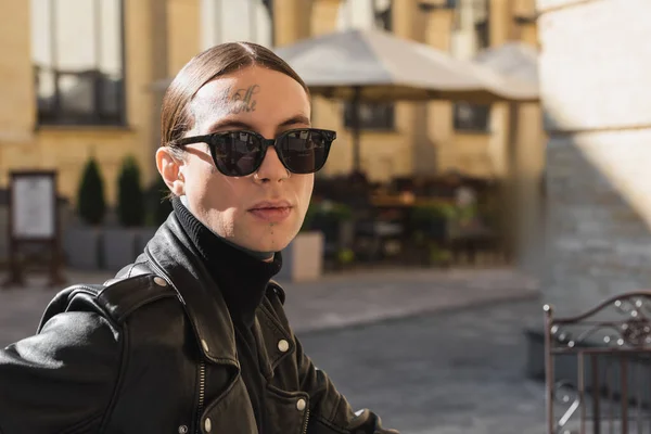 Tattooed young man in stylish sunglasses and black leather jacket looking at camera outdoors — Stockfoto