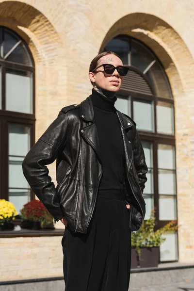 Tattooed man in stylish sunglasses and black leather jacket standing with hands in pockets on urban street — Stock Photo