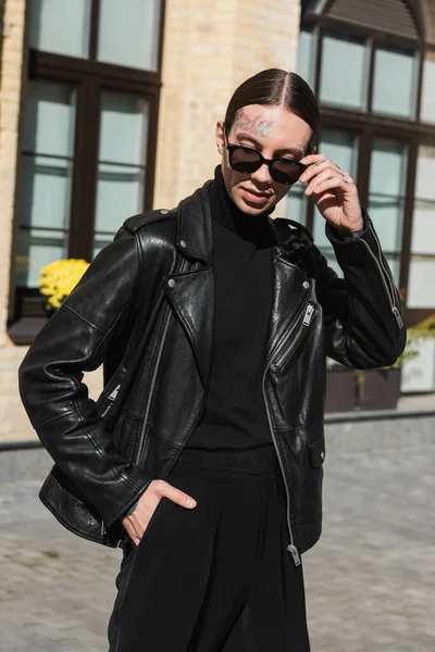 Tattooed young man adjusting stylish sunglasses and standing with hand in pocket on urban street — Stock Photo