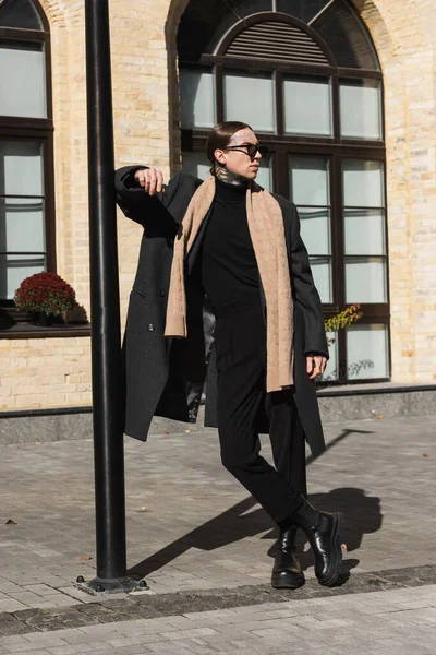 Full length of tattooed man in trendy coat and stylish sunglasses standing with crossed legs on urban street — Stock Photo