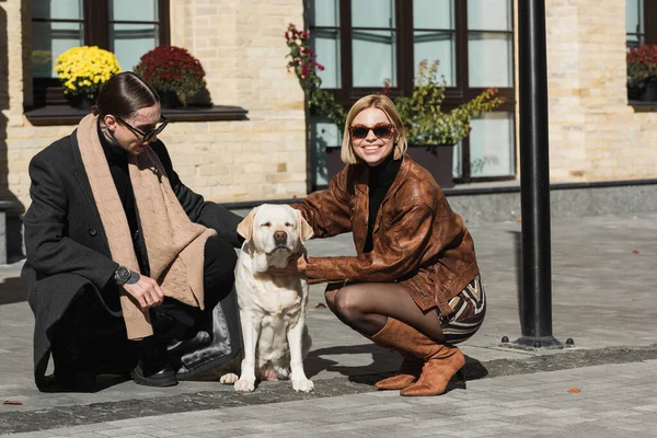 Full length of happy young couple in stylish sunglasses cuddling labrador on street — Photo de stock