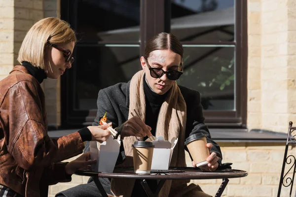 Stylish young couple eating asian food from carton boxes near coffee to go on bistro table — Photo de stock
