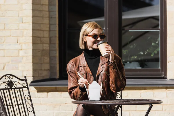 Felice donna bionda in occhiali da sole in possesso di bacchette vicino scatola da asporto con cibo asiatico e bere caffè — Foto stock