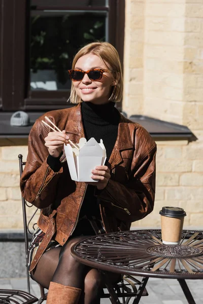 Donna felice con gli occhiali da sole che tiene le bacchette vicino alla scatola di cartone con cibo asiatico e tazza di carta sul tavolo da bistrot — Foto stock