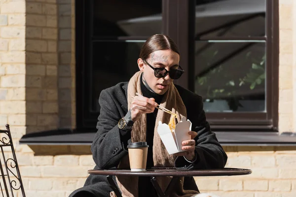 Tätowierter Mann mit Sonnenbrille und Mantel, der Essstäbchen hält, während er asiatisches Essen in der Nähe von Pappbechern auf dem Bistrotisch isst — Stockfoto