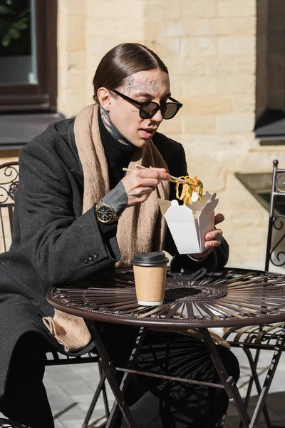 Tätowierter Mann mit stylischer Sonnenbrille, der Essstäbchen hält, während er asiatisches Essen in der Nähe von Pappbechern auf dem Bistrotisch isst — Stockfoto