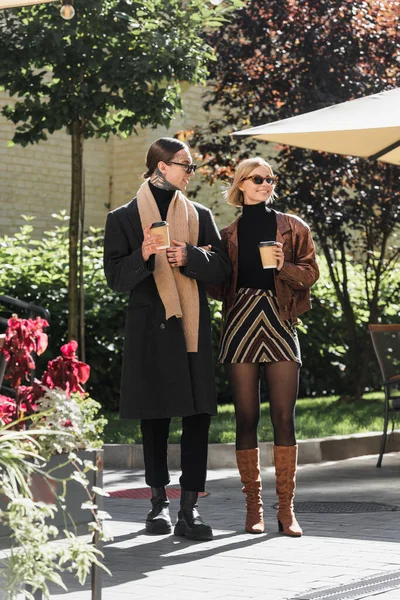 Full length of stylish couple in sunglasses holding paper cups while standing near outdoor cafe — Photo de stock
