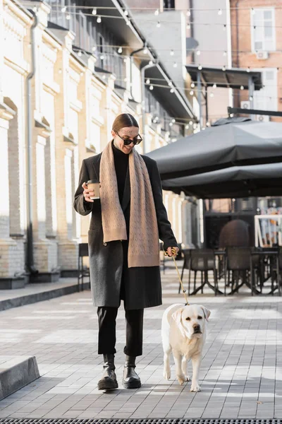 Full length of tattooed man in trendy sunglasses smiling and holding coffee to go while walking with labrador — Photo de stock
