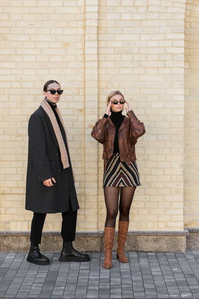 Full length of stylish woman adjusting sunglasses and standing near brick wall with tattooed man — Stockfoto