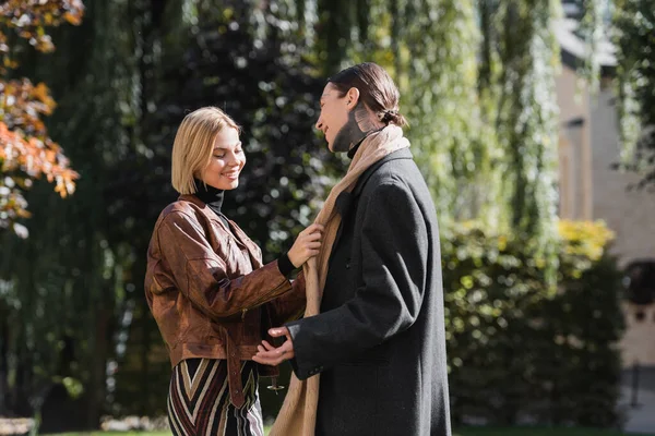 Happy blonde woman adjusting scarf of tattooed boyfriend during date in park — Stock Photo
