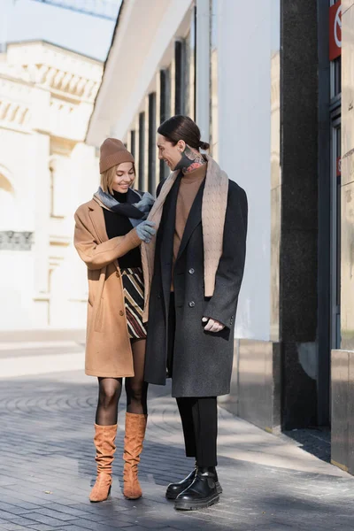 Full length of happy woman in hat adjusting scarf of man in coat — Stock Photo