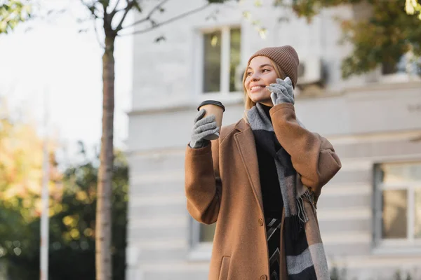 Smiling woman in hat and coat holding paper cup while talking on smartphone — Photo de stock