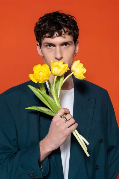 Trendy brunette man in blue blazer holding yellow tulips isolated on red - foto de stock