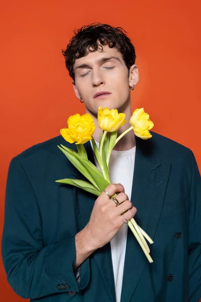 Brunette man in jacket closing eyes and holding yellow tulips isolated on red - foto de stock